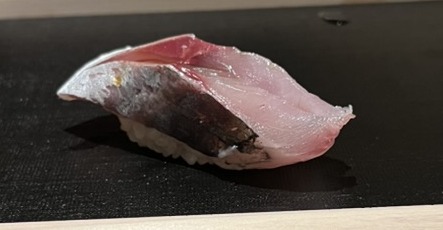 A piece of aji (horse mackerel) nigiri on a black board atop a wooden sushi box