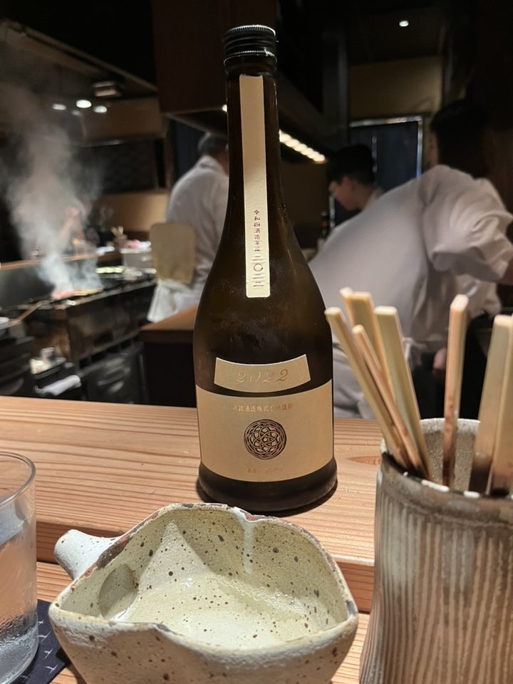 An amber Aramasa bottle, a 2022 special bottling with a spirograph crest, on a counter with a sake cup