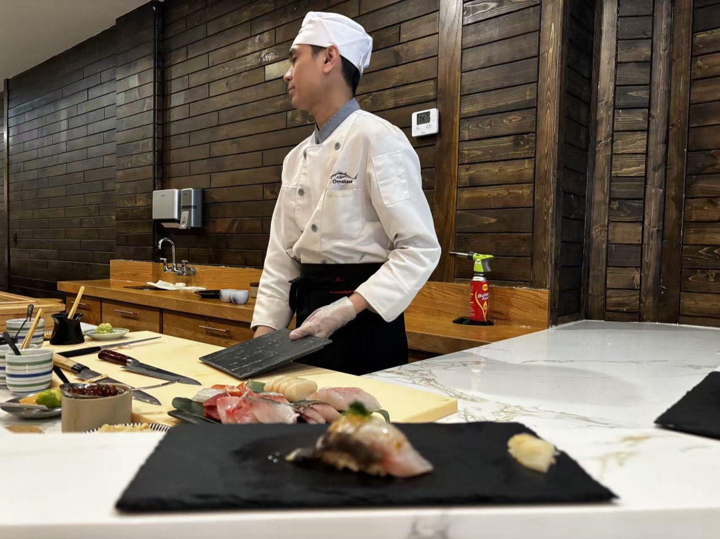 A cook in a tall white hat plating sushi at a New York counter while wearing a plastic glove, with a kitchen blowtorch and a Coca-Cola bottle on the counter behind him