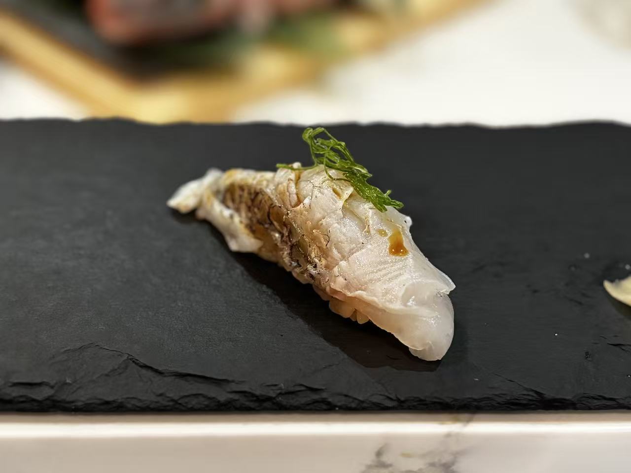 A single piece of white-fish nigiri on a black slate plate, topped with a small pinch of green garnish