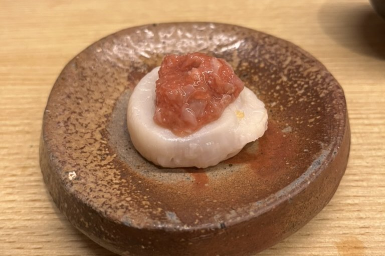 A grilled scallop topped with red shiokara on a rustic ceramic plate