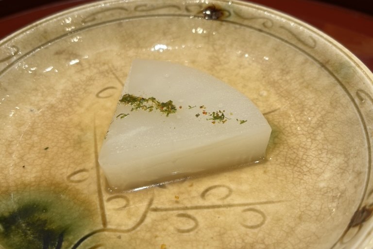 A wedge of simmered daikon in a pale dashi, dusted with yuzu, in a cream crackle-glazed bowl
