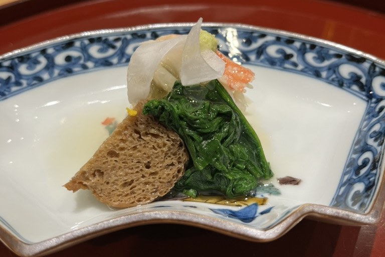 A composed opening dish of blanched greens, crab and shaved radish in a light dashi on a fan-shaped blue-and-white plate