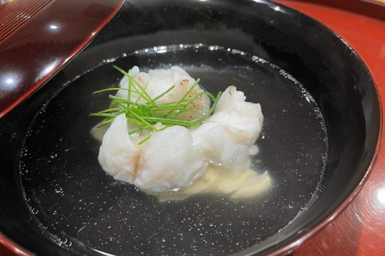A piece of lightly grilled white kue fish in a clear dashi with chive threads, in a black lacquer bowl