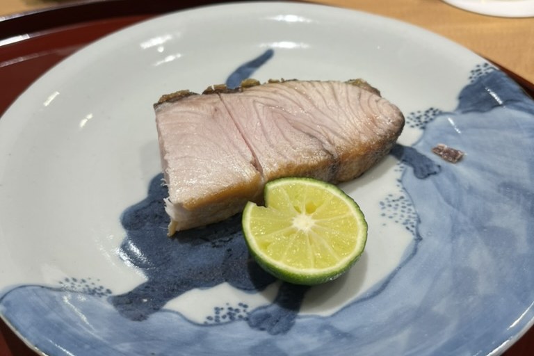 A thick, fatty grilled cut of sawara with a charred edge and a half of sudachi on a blue-and-white plate