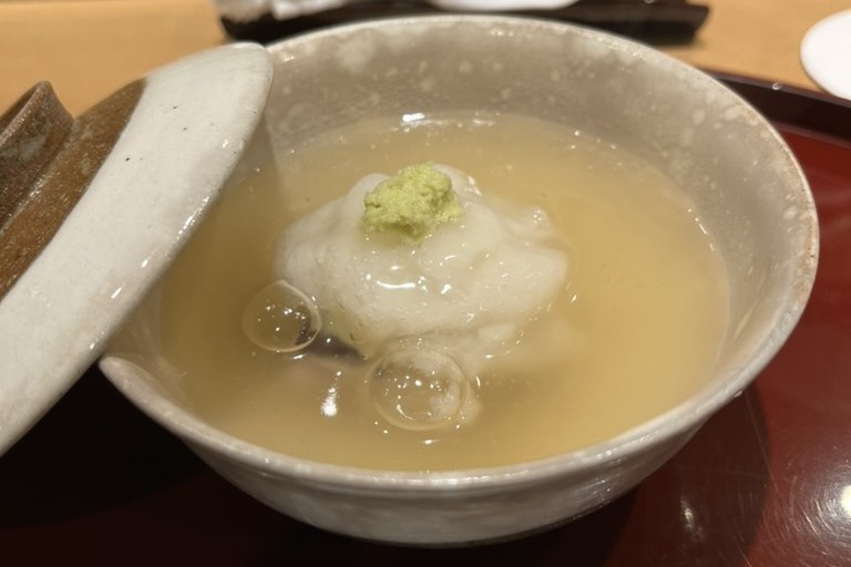 A soft eel shinjo dumpling topped with wasabi in a cloudy savoury broth, in a lidded ceramic bowl