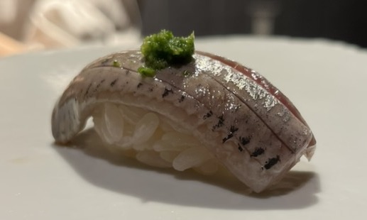 A piece of iwashi (sardine) nigiri topped with grated ginger and scallion on a white plate