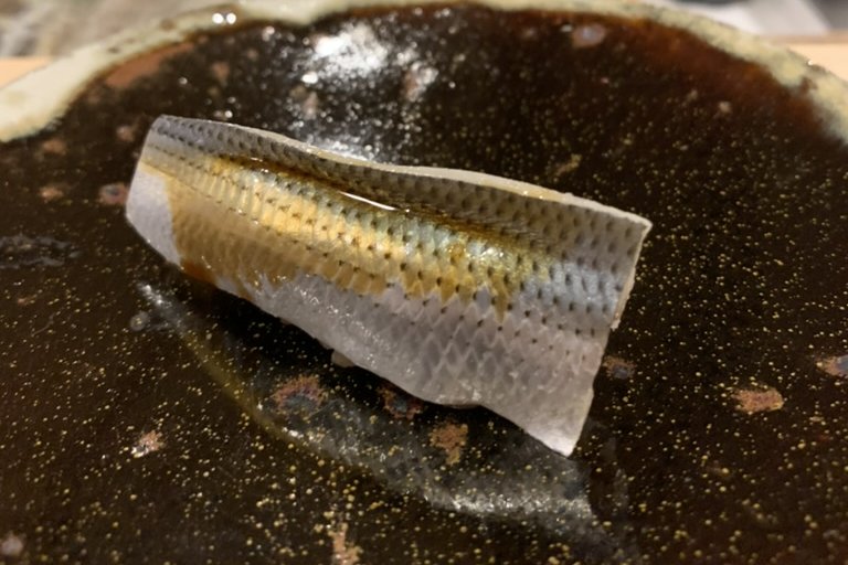 A kohada nigiri with finely scored silver skin on a dark gold-flecked plate, the neta box behind