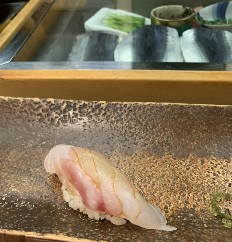 A piece of white-fish nigiri on a stone ledge in front of a glass-covered neta-case displaying the day's fish