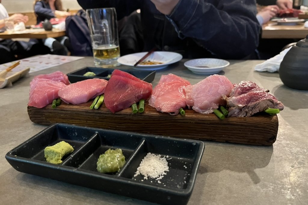 A long wooden board lined with different cuts of tuna from lean to fatty, with a tray of wasabi and salt