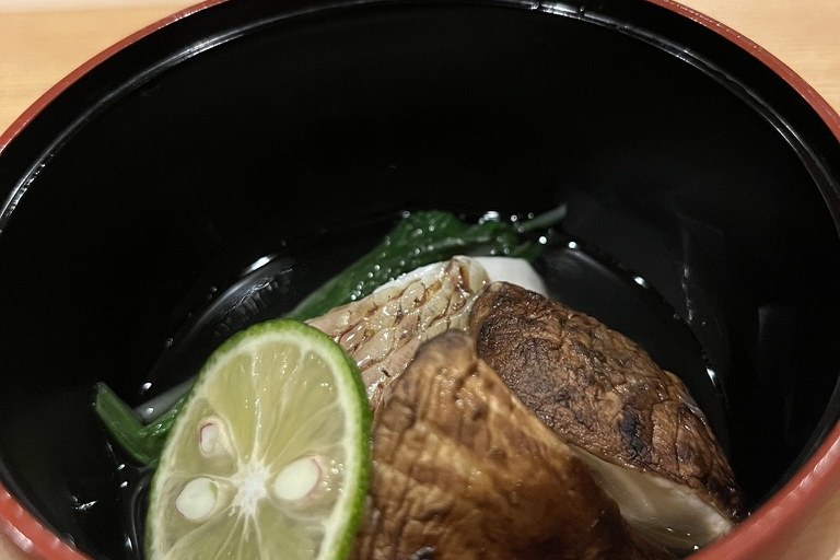 A red lacquer bowl of clear dashi with grilled matsutake mushroom, greens and a slice of sudachi