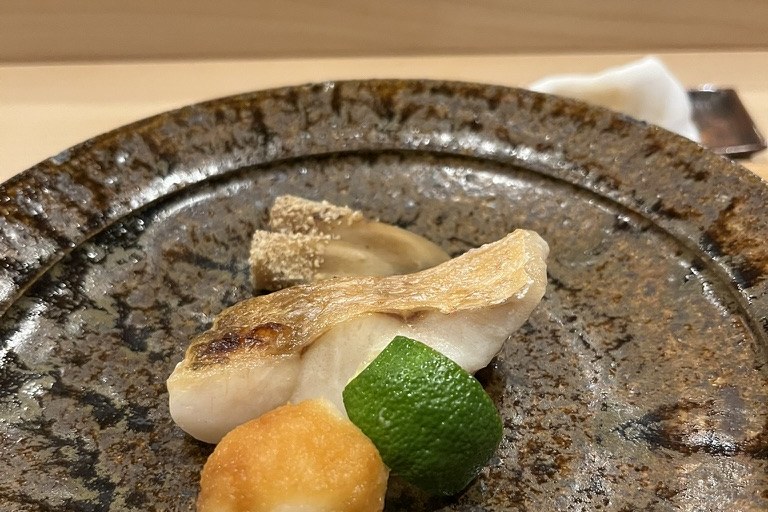 A piece of grilled nodoguro with crisped skin and a half of sudachi on a rustic dark plate