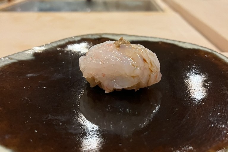 A mound of translucent shiroebi white shrimp formed into a nigiri on a glossy dark plate