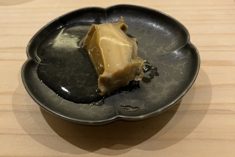 A piece of pale steamed abalone in a dark sauce on a black flower-shaped plate