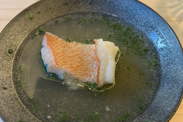 A piece of steamed golden-eye snapper in a clear broth scattered with chives, in a dark bowl