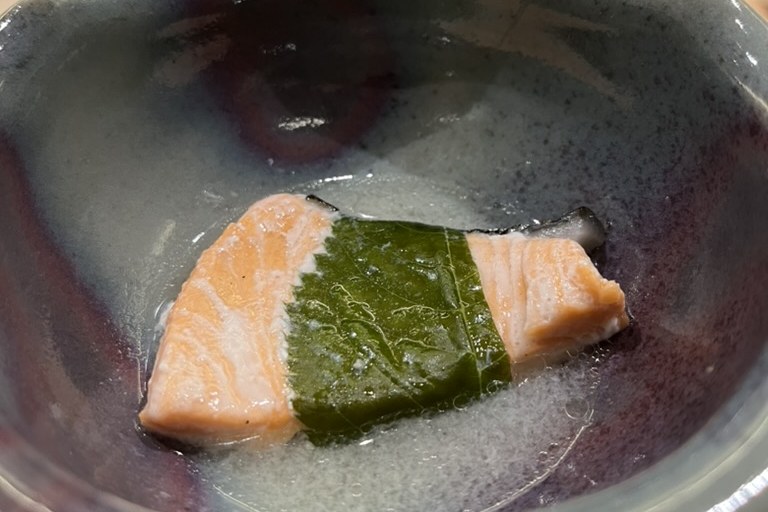 Two pieces of steamed trout flanking a sakura leaf in a clear broth, in a purple-glazed bowl
