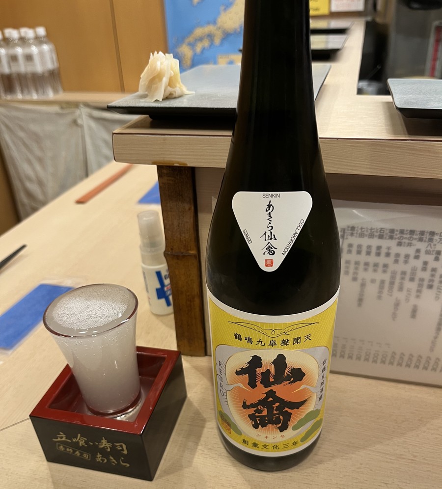 A glass of cloudy white nigori sake beside its bottle at a sushi counter