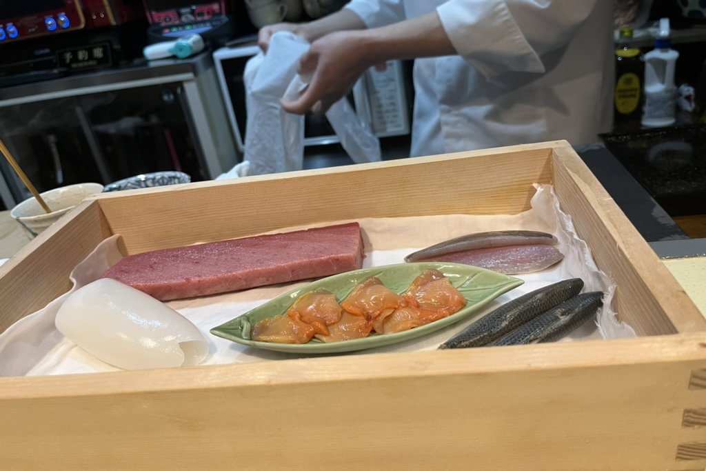 A wooden neta box at a sushi counter holding the day's prepared fish — a block of tuna, white squid, a plate of dressed ark shell, and silver-skinned fish — with the chef behind