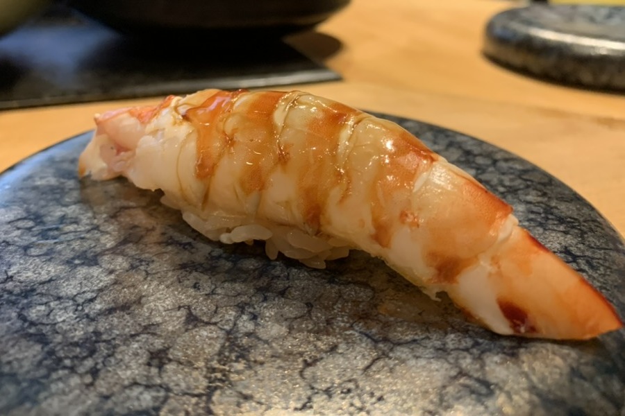 A kuruma-ebi nigiri glazed with nikiri, the prawn striped orange and white, on a speckled blue-grey plate