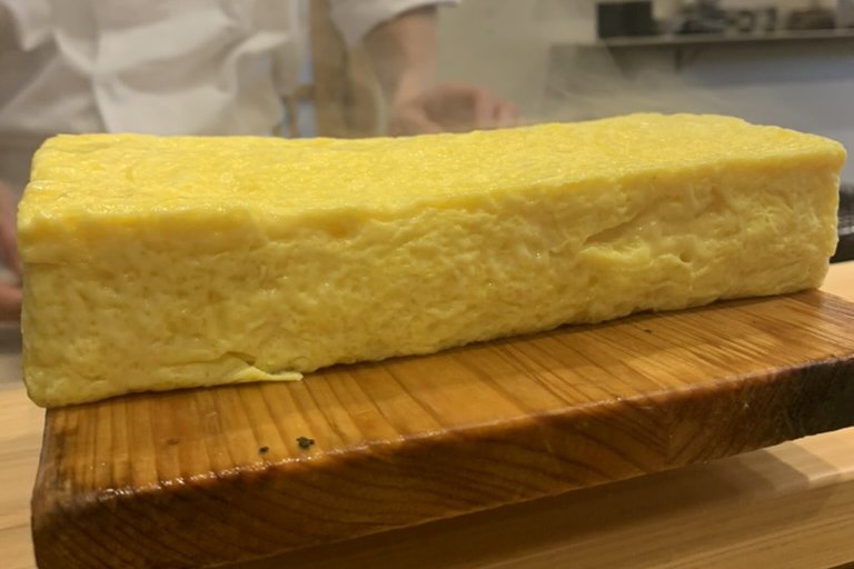A large, soft, pale-yellow rolled tamago loaf on a wooden board, the chef behind the counter