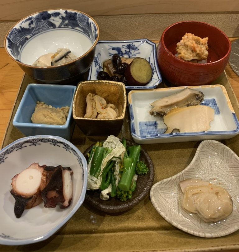 A tray of nine small tsumami dishes in assorted bowls and plates: simmered octopus, abalone, dressed shellfish, ohitashi greens and more