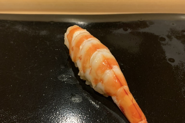 A whole kuruma-ebi (tiger prawn) nigiri, tail on, on a black plate