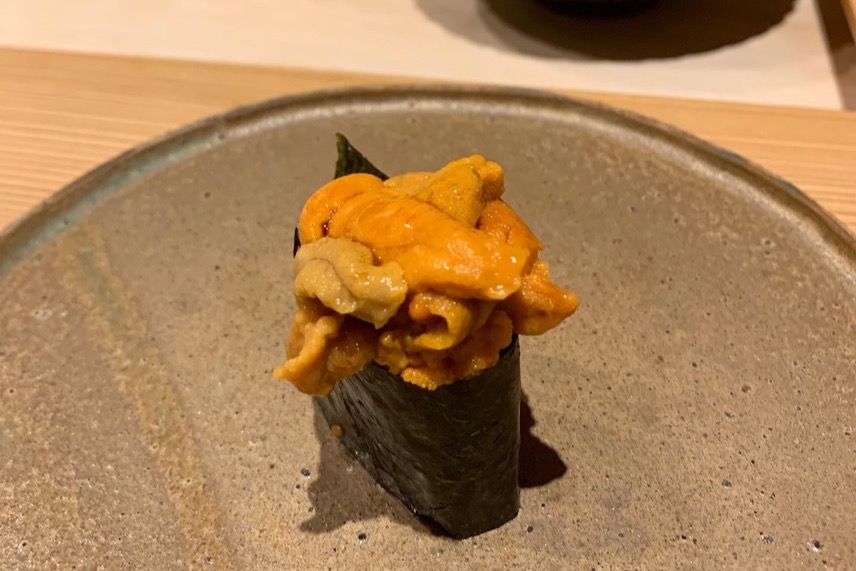 A uni gunkan wrapped in nori, topped with a mound of orange sea urchin, on a stone plate
