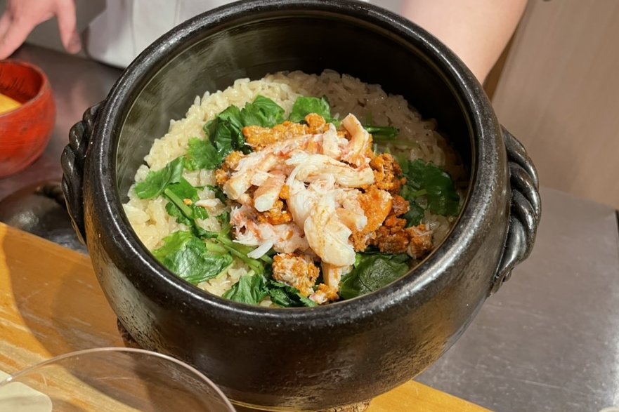 A clay pot of rice with crab meat, crab miso and greens, held at the counter