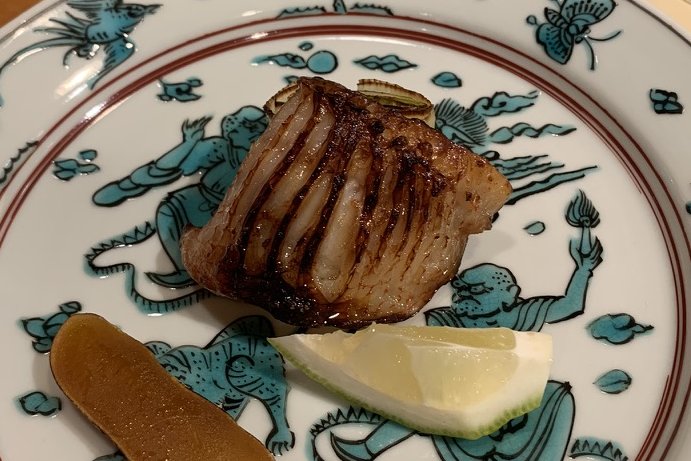 A roasted fish topped with karasumi on a blue-and-white kutani plate with a citrus wedge