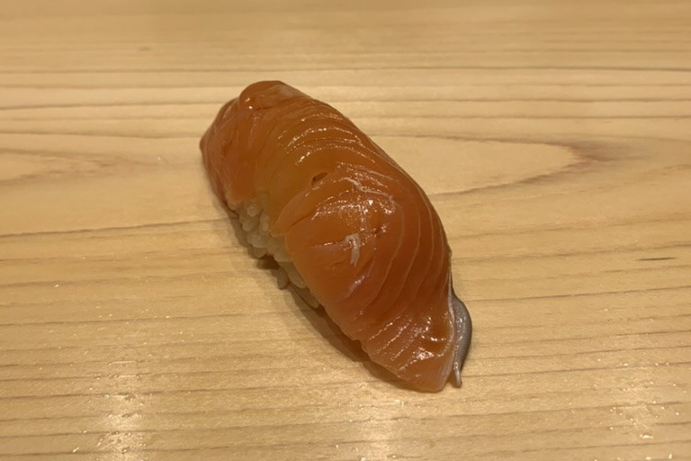 A single sakura-masu nigiri, its flesh a pale delicate orange, on a hinoki wood counter