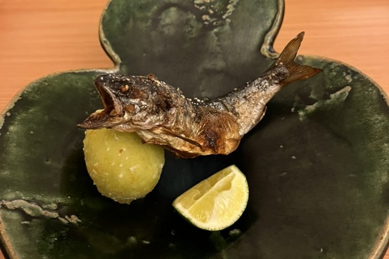 A salt-grilled ayu sweetfish curved on a green leaf-shaped plate, with a fried morsel and a wedge of sudachi