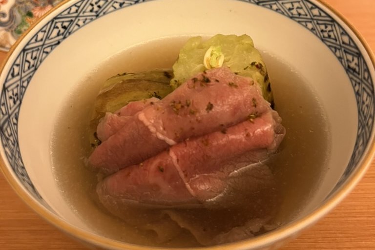 Thin slices of rare pink beef draped over a braised eggplant in a clear broth, in a blue-and-white bowl