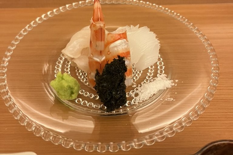 Sashimi on a beaded glass plate — a raw prawn upright beside thin-sliced squid, with wasabi, salt, and a dab of seaweed