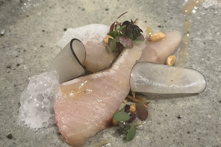 A French-style plated sawara dish — cured slices with microgreens, foam, puffed grains and shaved turnip on a grey plate