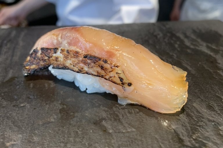 A lightly seared sawara (Spanish mackerel) nigiri, the skin charred, on a dark stone plate