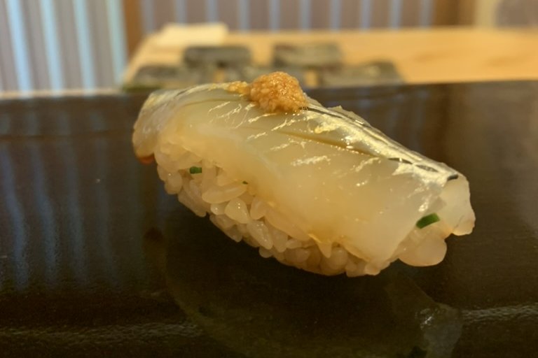 A translucent sayori (halfbeak) nigiri with a silver-blue stripe, topped with grated ginger, on a black plate
