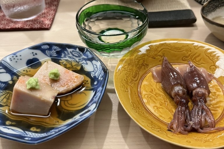 A slice of ankimo topped with wasabi on a blue-and-white plate, beside a green glass sake cup and a yellow dragon plate of small whole squid