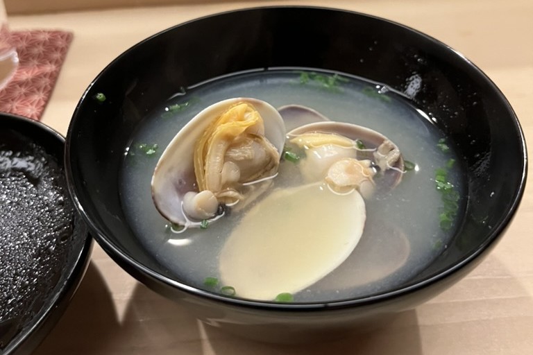 A black lacquer bowl of pale clear broth with two opened clams, chopped chives, and a smooth pale dumpling