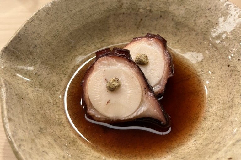 Two thick slices of slow-cooked octopus with pale cross-sections, sitting in dark tare in a speckled ceramic bowl