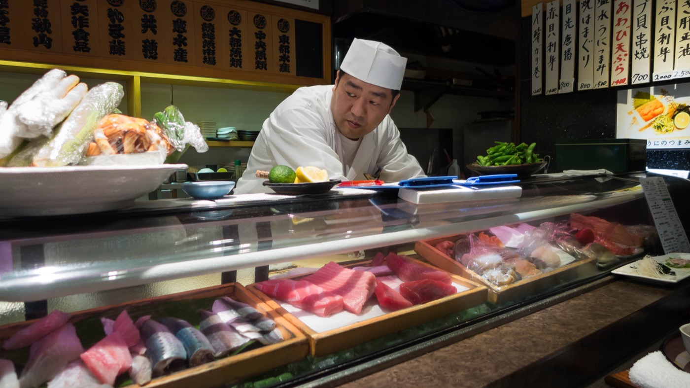 A sushi chef standing behind the neta case of fresh fish at his counter