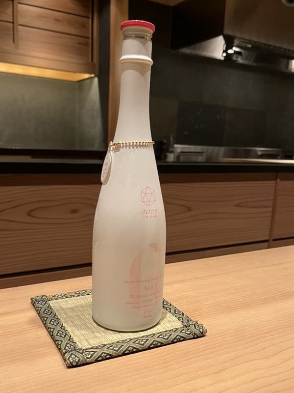 A frosted white bottle of Aramasa No.6 sake on a woven coaster on a hinoki sushi counter