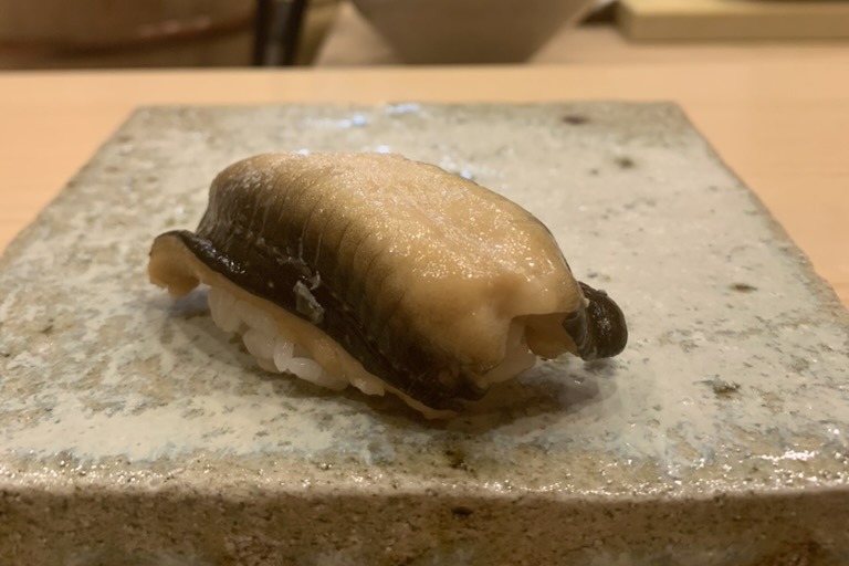 A simmered anago nigiri, the pale glazed eel folded over a small mound of rice with its dark skin visible at the edge, on a speckled stone slab