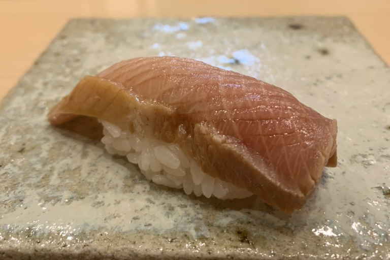 A buri nigiri, the pale pink yellowtail glazed with nikiri and hanging well past both ends of the rice, on a speckled stone slab