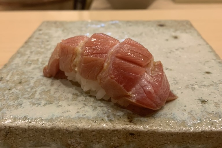 An otoro nigiri, the fatty tuna scored in a lattice and draped over a small mound of rice, on a speckled stone slab with the chef in whites behind