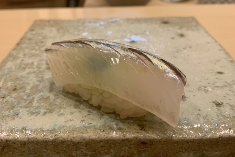 A sayori nigiri, the translucent halfbeak with a bright silver stripe along its edge, draped over rice on a speckled stone slab