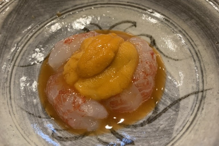 Peeled striped shrimp arranged in a ring with a mound of orange uni in the centre, sitting in a pale amber sauce in a brushed grey ceramic bowl