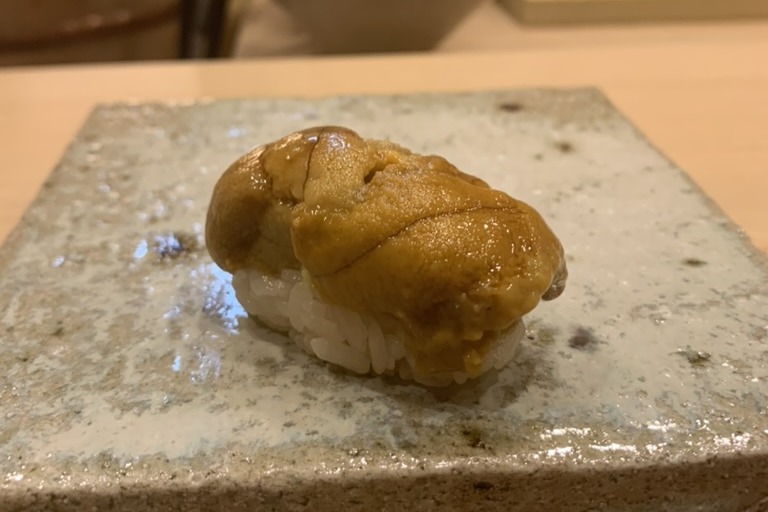 A uni nigiri, several tongues of orange sea urchin laid directly over rice without a nori wrap, on a speckled stone slab