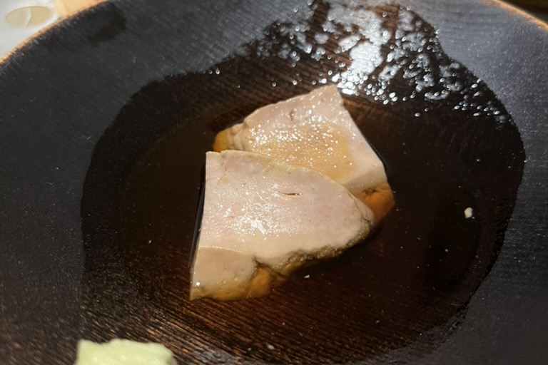 Two pieces of ankimo, monkfish liver, in a dark broth on a black lacquer plate with a dab of wasabi
