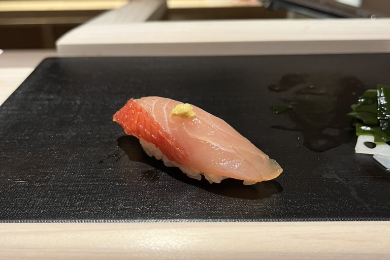 A single piece of kinmedai (golden eye snapper) nigiri on a black slate board at the counter
