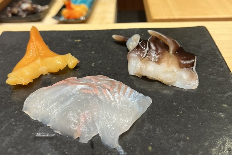 An opening assortment on slate — thin-sliced white fish, a shellfish, and a marinated bite